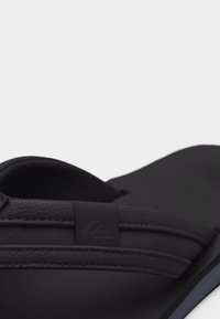 Κοντινή λήψη μαύρης σαγιονάρας με υφασμένους ιμάντες και μικρή ορθογώνια ετικέτα λογότυπου στον ιμάντα ανάμεσα στα δάχτυλα.