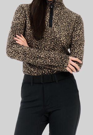 Frau mit langen dunklen Haaren, die einen Pullover mit Leopardenmuster und Reißverschluss bis zur Brust sowie eine schwarze Hose trägt, mit verschränkten Armen vor einem grauen Hintergrund steht.