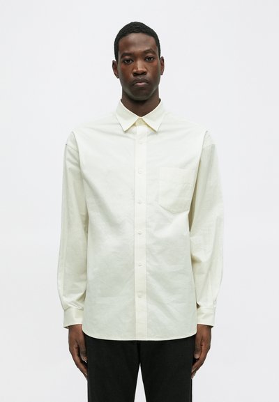 Jasna kremowa koszula z długim rękawem z miękkiej bawełny. Posiada kołnierz zapinany na guziki, pięć guzików oraz kieszonkę na lewej piersi. Zmięta faktura.