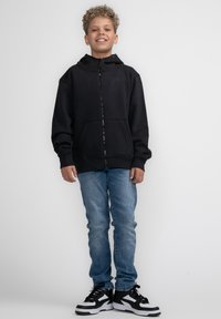 Felpa nera con cerniera e tasca anteriore, indossata con jeans azzurri chiari e sneakers bianco e nere. Il modello ha capelli ricci.