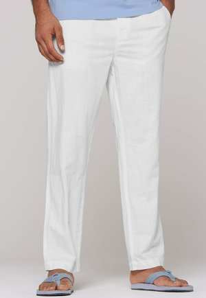 Mann trägt weiße Leinenhose, blaues T-Shirt und hellblaue Sandalen, steht mit den Händen in den Taschen vor einem einfarbigen Hintergrund.