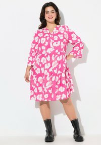 Kleid mit floralem Muster in leuchtendem Pink und Weiß, knielang, mit gestuftem Design und Dreiviertelärmeln; kombiniert mit schwarzen Stiefeletten.