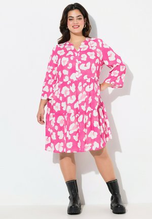 Vestido de patrón floral en rosa brillante y blanco, hasta la rodilla, con un diseño en capas y mangas tres cuartos; combinado con botas negras hasta el tobillo.