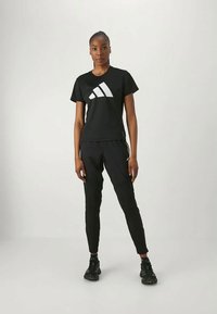 Musta urheiluasu, jossa on lyhythihainen t-paita valkoisella Adidas-logolla ja siihen sopivat kapenevat housut. Tekstuurin ilme on sileä ja hyvin istuva.