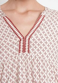 Tunique blanche avec motif losange rouge, encolure en V avec broderie en zigzag le long du col, corsage froncé, texture de tissu léger.