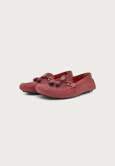Paire de mocassins en daim rouge avec pompons décoratifs, coutures apparentes et semelles en caoutchouc à picots sur fond blanc.