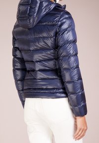 Giacca a vento blu navy con cappuccio, caratterizzata da sezioni trapuntate orizzontali, texture lucida e polsini elasticizzati. La vista posteriore mostra una silhouette aderente.