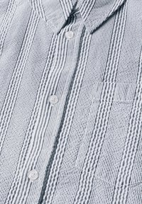 Modré a bílé pruhované tričko s krátkým rukávem, s límečkem na knoflíky, malou náprsní kapsou a texturovanou tkaninou s vyvýšenými horizontálními liniemi.