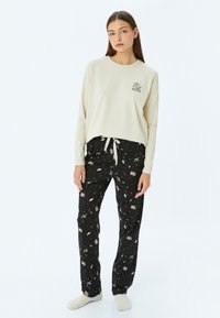 Beige lange mouwen top met de tekst "Magic"; zwarte broek met een speels patroon van muzieknoten en voorwerpen, voorzien van een trekkoord in de taille.