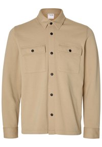 Veste-chemise beige à manches longues avec boutons-pression noirs, deux poches poitrine à rabat à l'avant et col pointu.