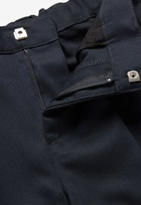 Gros plan sur un pantalon bleu foncé montrant une fermeture éclair partiellement ouverte et une fermeture à crochet et œil en métal sur une ceinture en tissu épais.