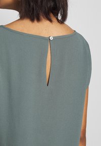 Retro di una persona che indossa un top testurizzato verde opaco con una piccola apertura a chiave fissata da un singolo bottone sul collo.