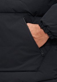 Czarna puchowa kurtka z ręką w bocznej kieszeni na zamek. Tkanina jest gładka o matowym wykończeniu, z marszczonym mankietem.