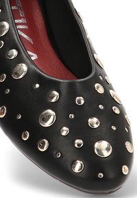 Zapato de cuero negro con puntera redonda, que presenta tachuelas metálicas de diferentes tamaños en la parte superior. Forro interior rojo, textura suave.