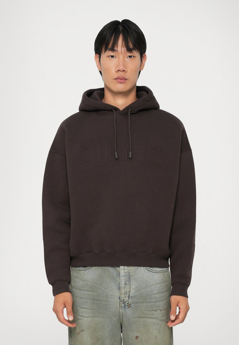 Felpa con cappuccio di un marrone scuro e vestibilità rilassata, con un logo goffrato discreto sulla parte anteriore e coulisse regolabili. Texture morbida.