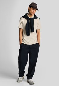 Polo beige, felpa blu navy appoggiata sulle spalle, pantaloni blu navy larghi, scarpe grigie e bianche, berretto scuro con logo.