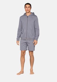 Man staat blootsvoets, gekleed in een bijpassend grijs capuchonvest met rits en shorts met een subtiel cirkelpatroon op een witte achtergrond.