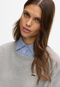 Γυναίκα με μακριά καστανά μαλλιά, φορώντας ένα γκρίζο φούτερ πάνω από ένα μπλε καρό πουκάμισο, κοιτάζοντας ελαφρώς στραμμένη προς την κάμερα.