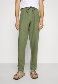 Person iført olivengrønne drawstring-bukser, hvid skjorte og beige ruskindssandaler med spænde, stående foran en ensfarvet lys baggrund.