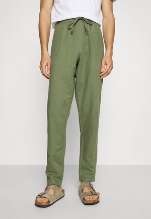 Persoon in olijfgroene broek met trekkoord, wit shirt en beige suède sandalen met gesps, staande tegen een effen lichte achtergrond.