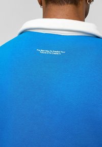 Blauwe polo met een witte kraag, gemaakt van een gestructureerde stof. Een motiverende quote is in witte tekst op de rug gedrukt.