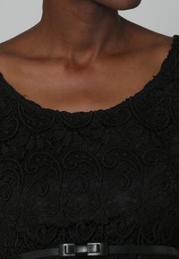 Czarna sukienka z koronką, z kwiatowym wzorem, okrągłym dekoltem i krótkimi rękawami. Czarny pasek podkreśla talię.