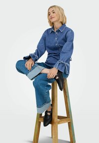 Džínová košile s knoflíkovým zapínáním vepředu, dlouhými rukávy se šnůrkami, spárovaná se širokými džínovými kalhotami s manžetou. Modelka má na sobě černé boty.