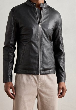 Homme portant une veste en cuir noire zippée avec un col pression et un pantalon beige, debout devant un fond clair uni.