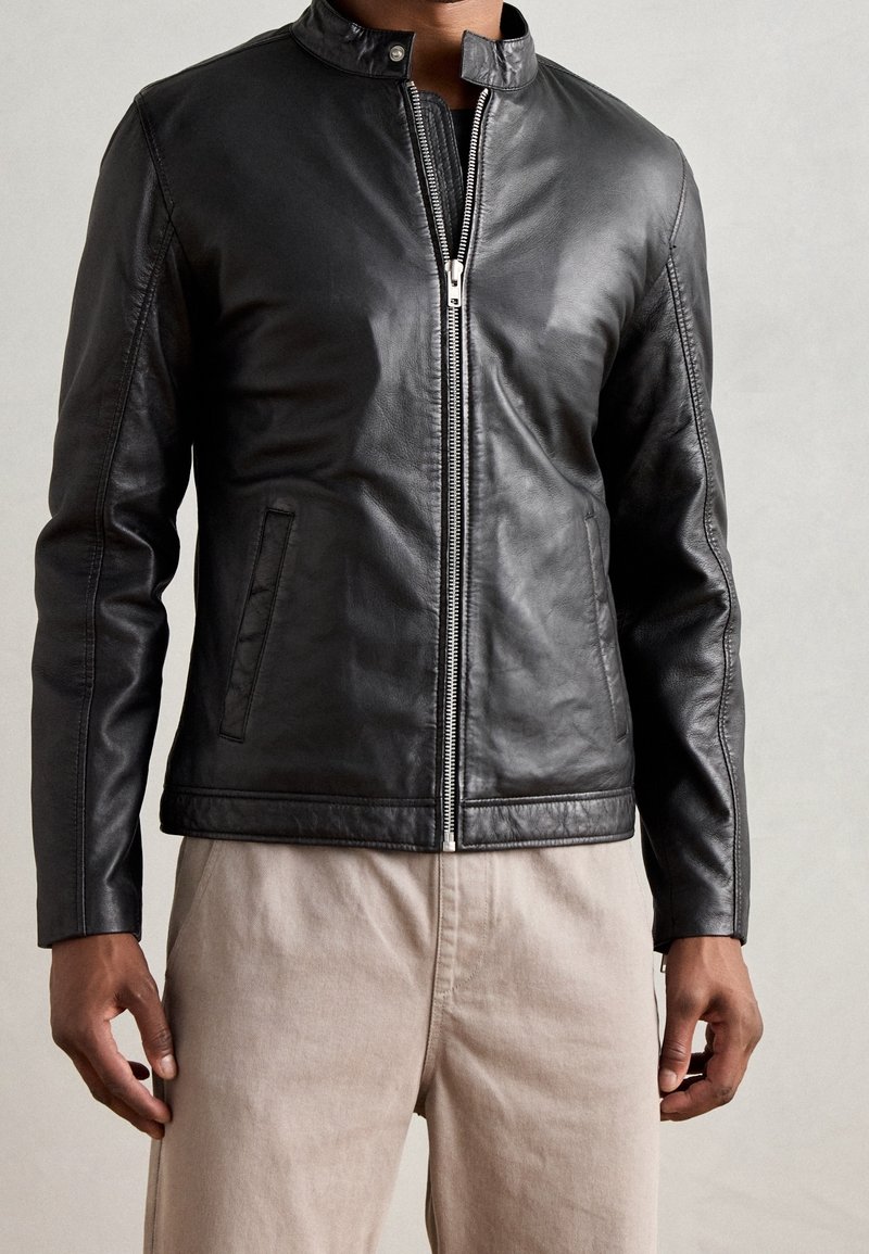 Homme portant une veste en cuir noire zippée avec un col pression et un pantalon beige, debout devant un fond clair uni.