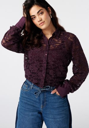 Jeune femme aux cheveux ondulés foncés portant un chemisier en dentelle violette transparente et un jean bleu taille haute avec une ceinture nouée, posant avec la main dans les cheveux.