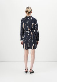 Femme aux cheveux blonds en chignon bas, portant une robe à motifs abstraits noirs, beiges et blancs, ainsi que des escarpins noirs à talons ouverts, se tenant face à un mur blanc.