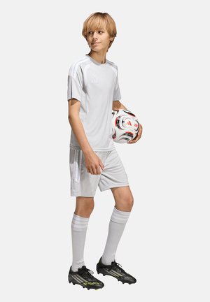 Young boy in white soccer uniform holding a soccer ball, standing and looking to the side against a plain background.