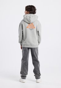 Felpa grigia con un grande grafico triangolare arancione etichettato "COLORADO TRAIL CLUB 1989" sulla schiena. Indossata con pantaloni grigi e scarpe da ginnastica bianche.