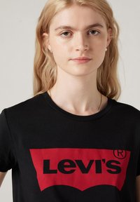 Sort bomulds-T-shirt med et stort rødt Levi's-logo over brystet, med rund halsudskæring og korte ærmer. Simpelt design.