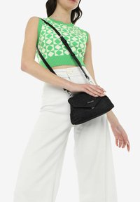 Sac en bandoulière noir avec tissu texturé, fermeture à rabat et quincaillerie argentée. Porté avec un crop top vert à motifs et un pantalon large blanc.