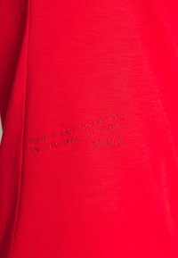 Tela roja con texto cosido que dice "NO DEJES QUE LO BUENO SE INTERPONGA EN EL CAMINO DE LO GRANDE PUMA" en una fuente pequeña.