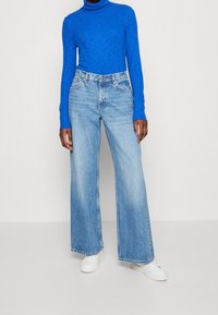 Maglione a collo alto blu con texture abbinato a jeans in denim blu chiaro a gamba larga e sneakers bianche. Outfit casual, dettagli visibili minimi.