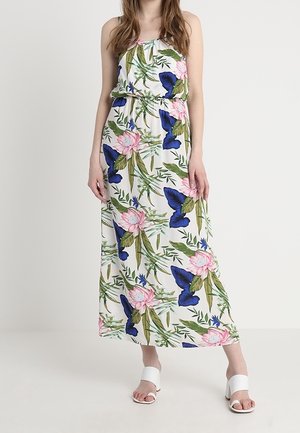 Femme portant une robe maxi blanche sans manches avec un imprimé floral et papillons bleu et rose, associée à des sandales blanches à talons blocs et ouvertures à l'avant.
