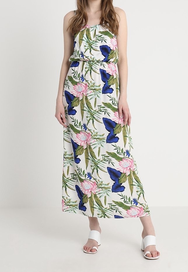 Femme portant une robe maxi blanche sans manches avec un imprimé floral et papillons bleu et rose, associée à des sandales blanches à talons blocs et ouvertures à l'avant.