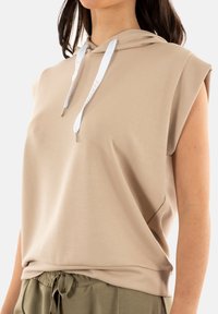 Sudadera sin mangas beige de suave tela, con bolsillo canguro en el frente y cordones blancos con puntas plateadas.