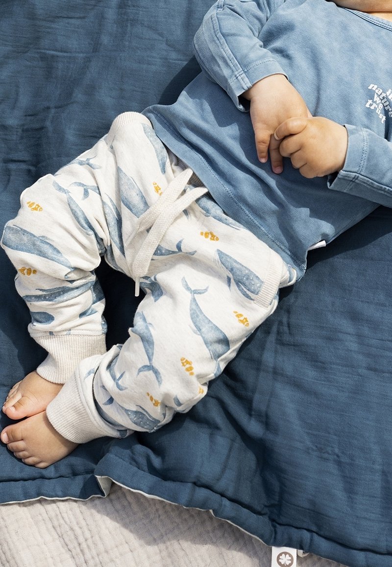 Enfant portant un t-shirt bleu à manches longues et un pantalon blanc avec des motifs de baleines et de poissons, allongé sur une couverture bleu foncé.