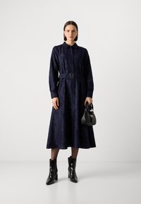 Donkerblauwe shirtjurk met bloemenpatroon, knoopsluiting, ceintuur in de taille, lange mouwen, gecombineerd met zwarte enkellaarsjes met hak en een kleine handtas.