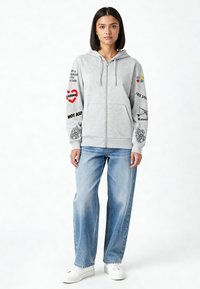 Felpa con cappuccio grigia con zip e maniche lunghe, con grafiche stampate tra cui cuori e testo. Abbinata a jeans azzurri e sneakers bianche.