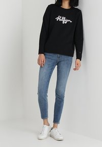 Czarna bluza z napisem "Hollister" w białym kolorze, zestawiona z jasnoniebieskimi obcisłymi dżinsami i białymi trampkami, na neutralnym tle.