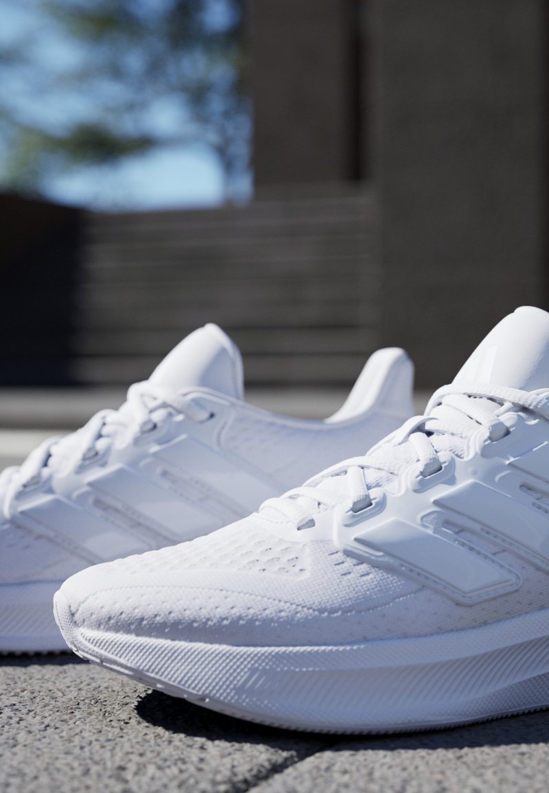 Paire de chaussures de course blanches placées sur une surface pavée extérieure en plein soleil, avec des marches floues et de la verdure en arrière-plan.