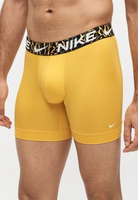 Torse et parties supérieures des jambes masculins portant un boxer jaune ajusté Nike avec ceinture noire et jaune à motif et logos blancs Nike swoosh.