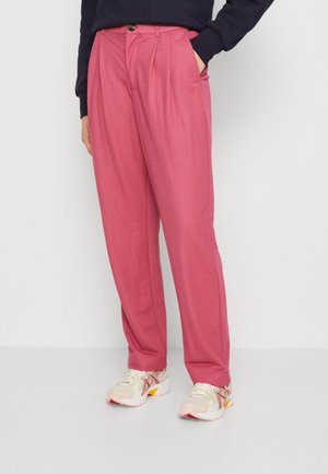 Person, der bærer løse lyserøde bukser med forreste læg, sidelommer, en mørk top og hvide sneakers med røde og gule detaljer.