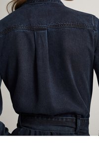Vue de dos d'une personne portant une veste en jean foncé rentrée dans un pantalon en jean taille haute assorti avec une ceinture.