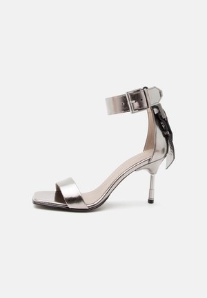 Strieborná metalická otvorená sandála na vysokom podpätku s remienkom okolo členka a prackou, zdobená ozdobnou mašľou na zadnej strane.