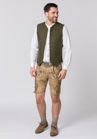 Gilet en laine vert avec des boutons en argent, chemise blanche en dessous, shorts en cuir beige avec des broderies vertes complexes, et chaussures marron.